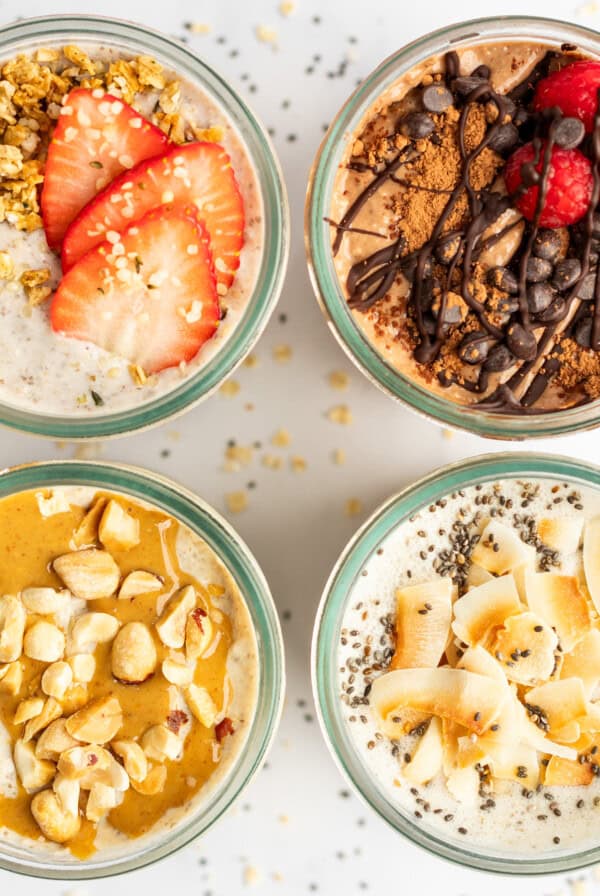 Top-down view of four jars filled with different flavors of overnight oats, garnished with toppings like strawberries, chocolate, raspberries, nuts, and coconut flakes, arranged in a 2x2 grid.