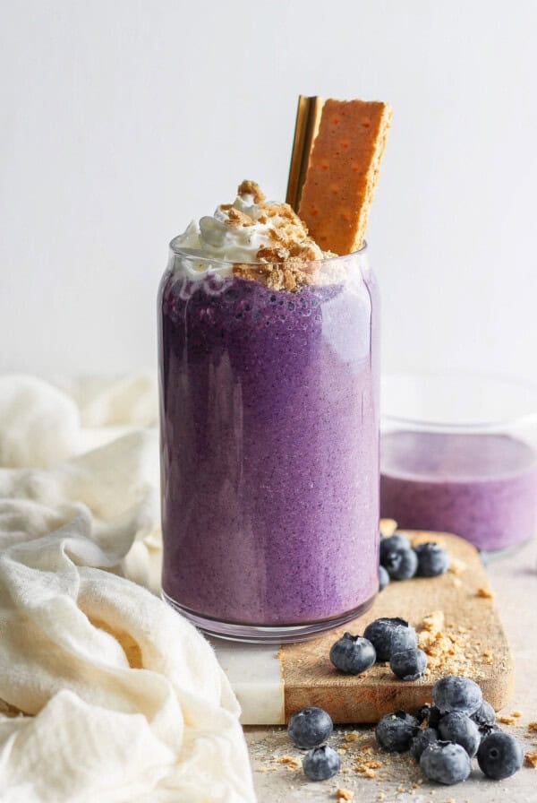 A vibrant purple smoothie topped with whipped cream and a cookie, garnished with blueberries on a light, textured background.