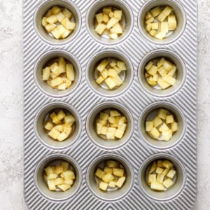 A muffin tin containing evenly chopped apples in each compartment, set on a light textured surface.