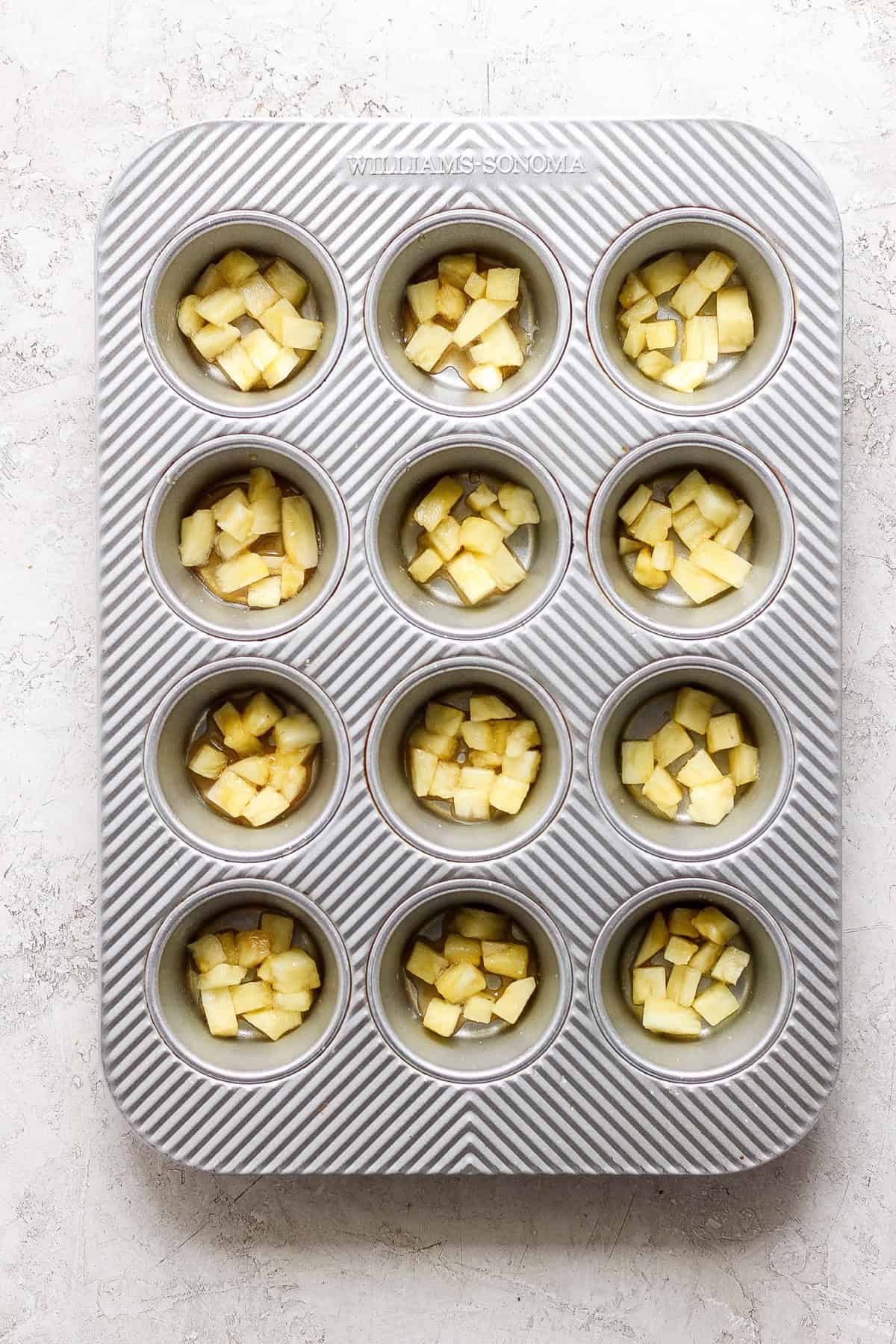 A muffin tin containing evenly chopped apples in each compartment, set on a light textured surface.