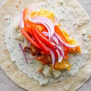 A flour tortilla spread with herb cream cheese topped with slices of red bell pepper, red onion, and chunks of chicken and orange.