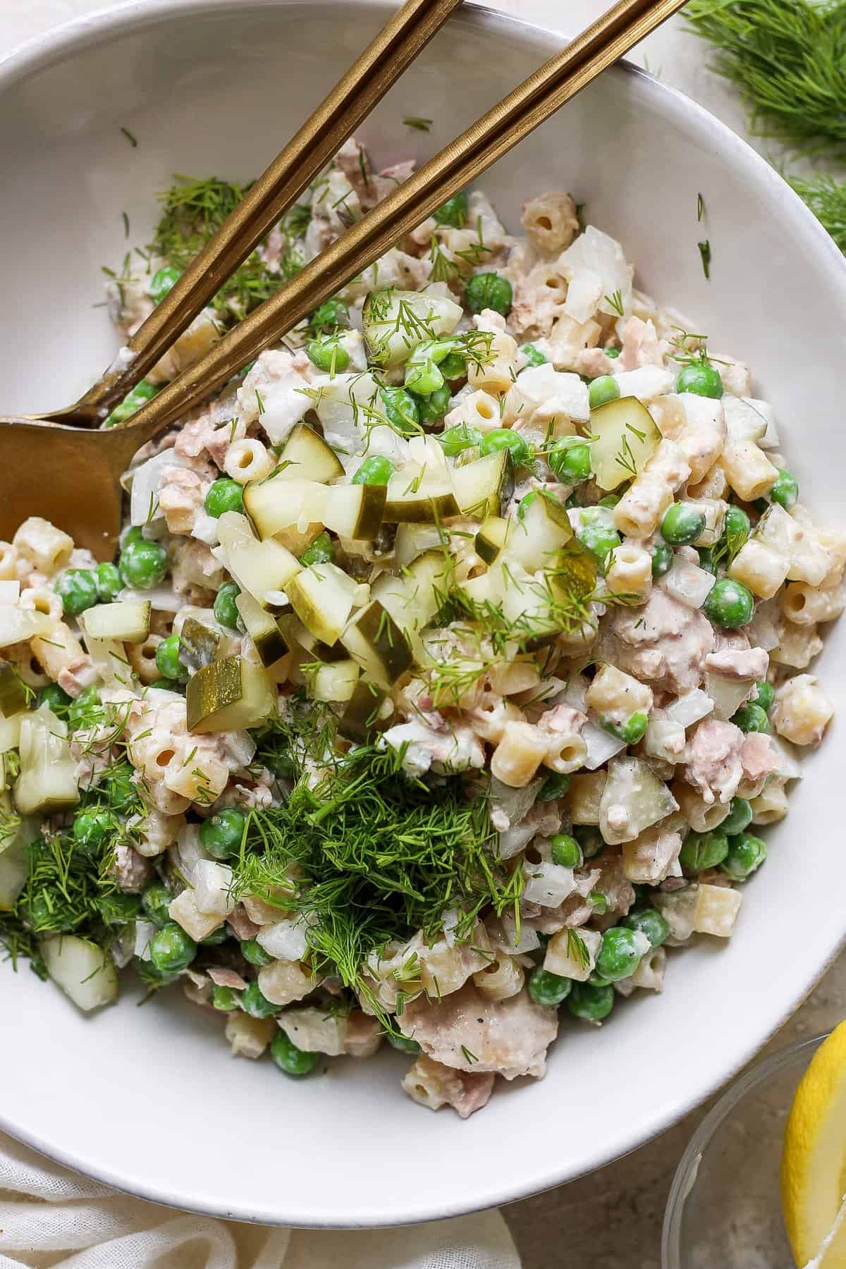 A bowl of tuna salad with peas, chopped cucumbers, and dill, served with a pair of gold chopsticks on a white plate.