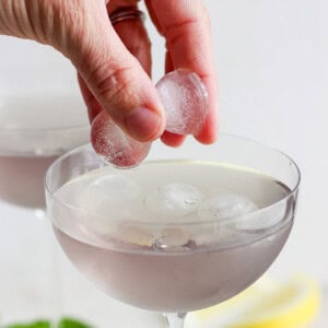 A hand placing ice cubes into a cocktail glass filled with a clear drink, with more ice-filled glasses and lemon slices blurred in the background.