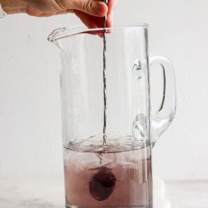 A hand uses a metal spoon to stir a dissolving purple effervescent tablet in a clear glass pitcher filled with water.