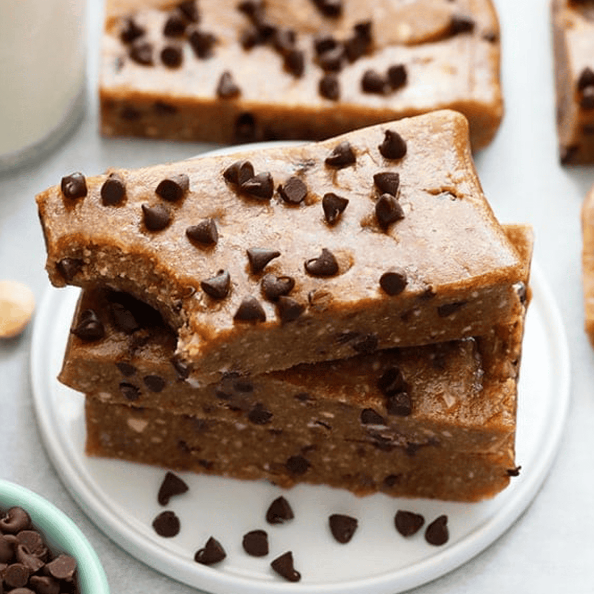 Three homemade cookie dough bars with chocolate chips stacked on a white round plate. One bar has a bite taken out of it, reminiscent of classic homemade Larabars. A small bowl with chocolate chips is visible in the foreground.