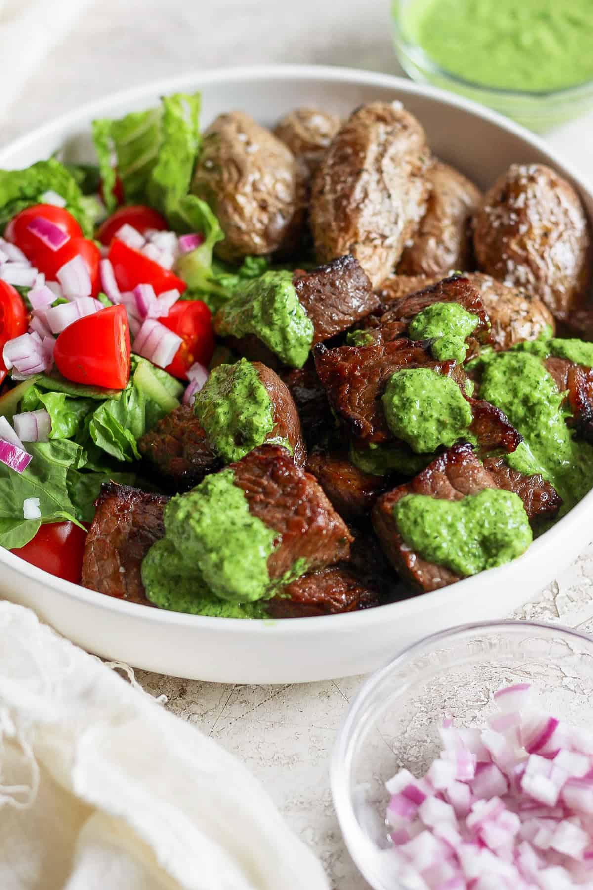 A bowl with grilled steak topped with green chimi-churri sauce, roasted potatoes, and a salad of lettuce, cherry tomatoes, and red onions. A small dish of chopped red onions is beside the bowl.