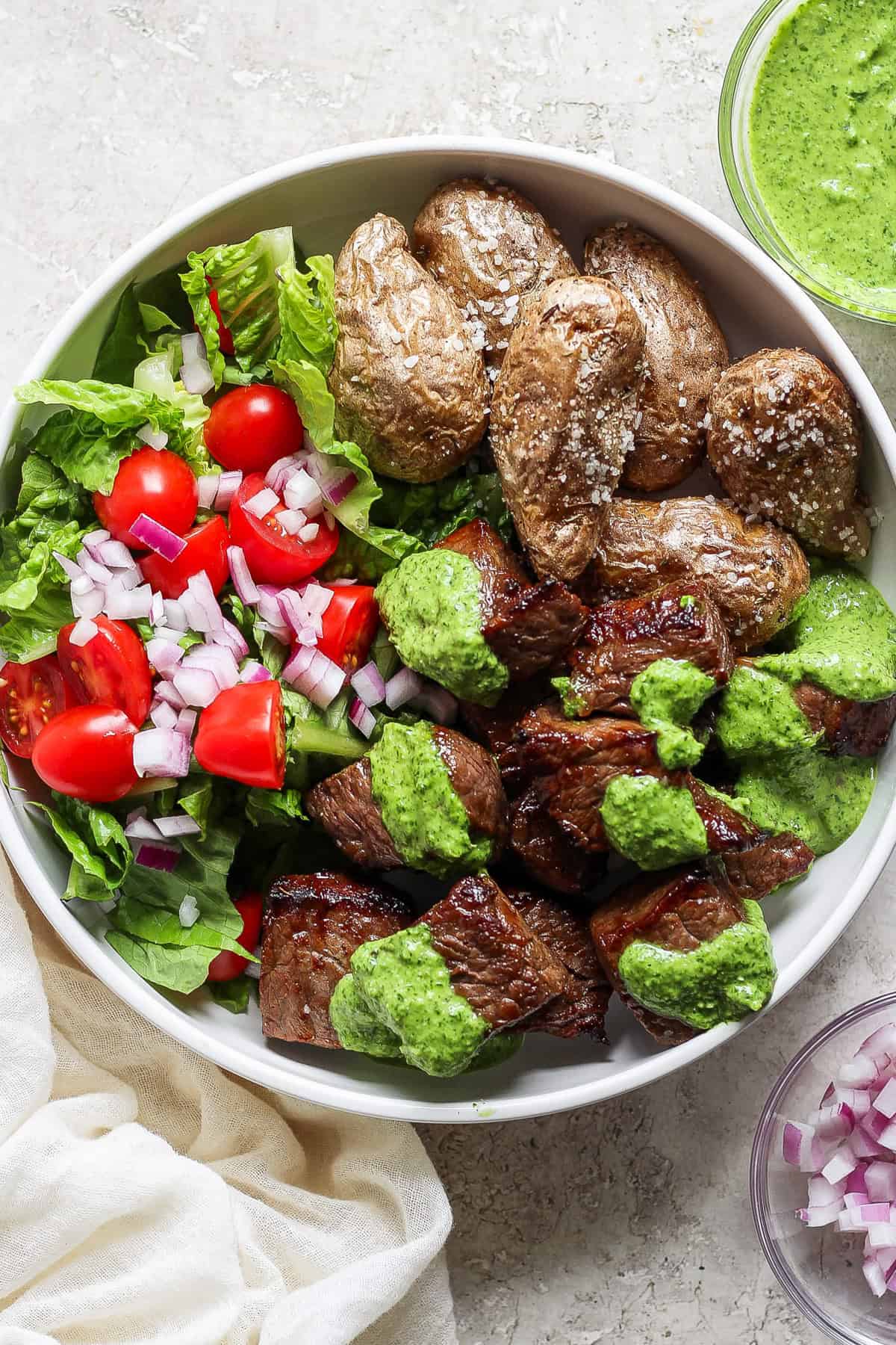 A bowl of grilled steak chunks with green sauce, roasted potatoes, and a side salad of lettuce, tomatoes, and chopped red onions.