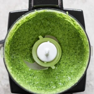 A top-down view of a blender with green sauce inside, featuring visible greens and herbs blended to a smooth consistency.