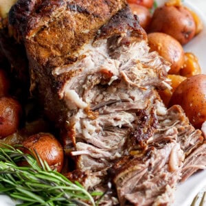 A close-up of a seasoned, cooked roast surrounded by whole red potatoes and fresh rosemary sprigs on a white platter. A fork and knife are set to the side.