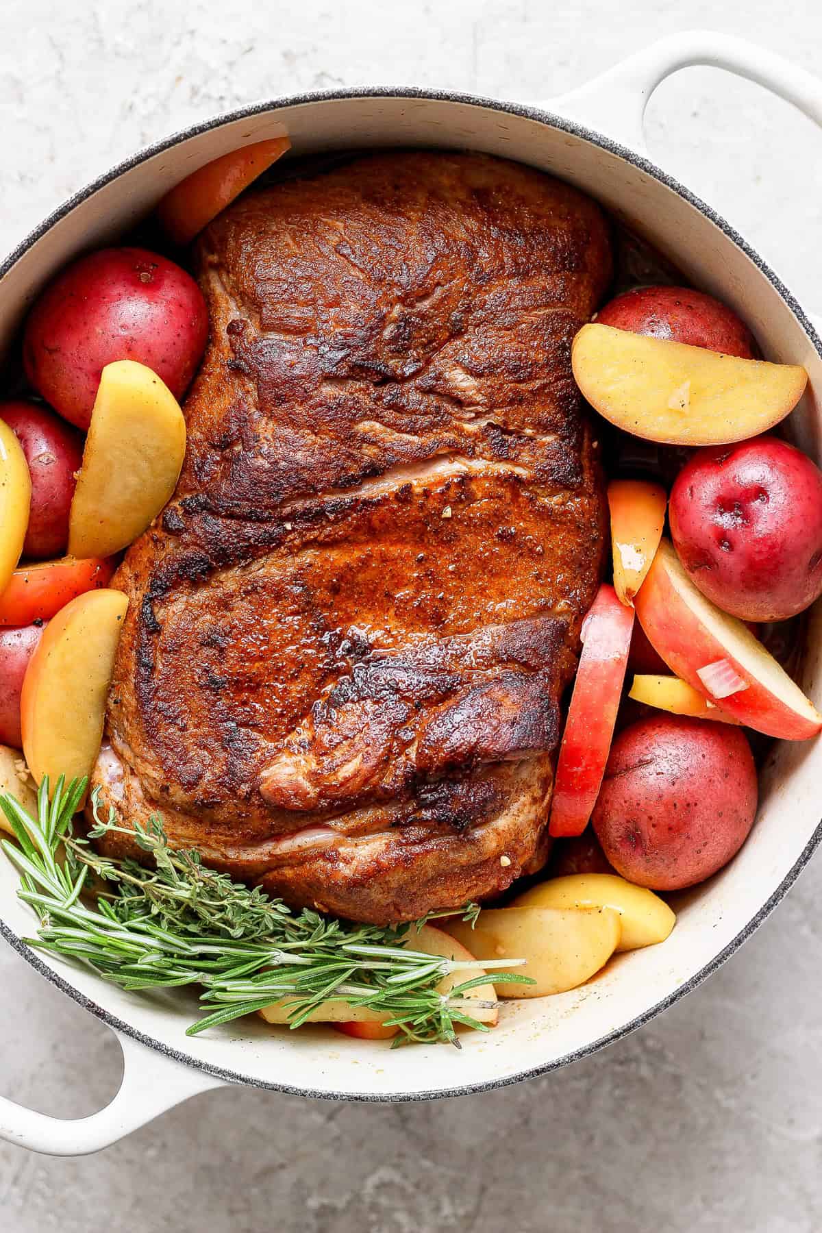 A cooked pork roast with a crispy exterior, surrounded by red potatoes, apple slices, and sprigs of rosemary in a white cast iron pot.