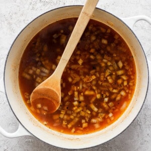 A white pot filled with a red broth containing chopped onions, stirred with a wooden spoon, placed on a gray textured surface.