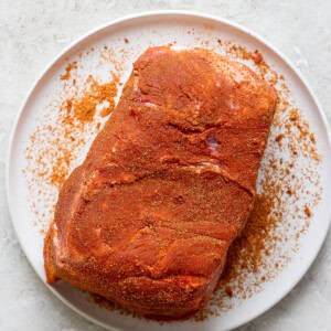 A piece of seasoned raw meat on a white plate, placed on a textured white surface.