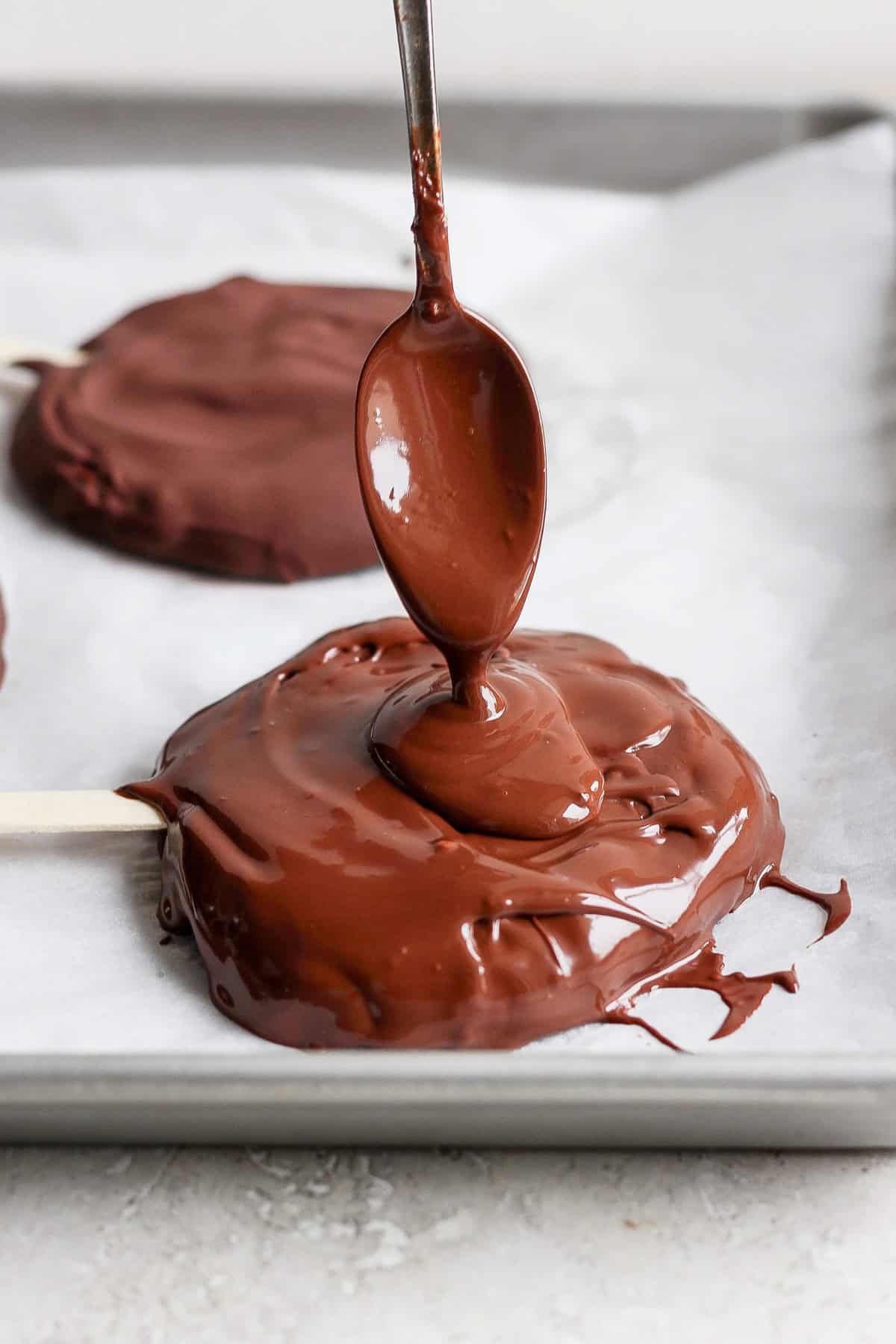 A spoon drizzles melted chocolate onto a round treat on a parchment-lined tray with a popsicle stick.