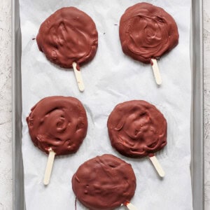 Five chocolate-covered treats on wooden sticks rest on a parchment-lined baking sheet.