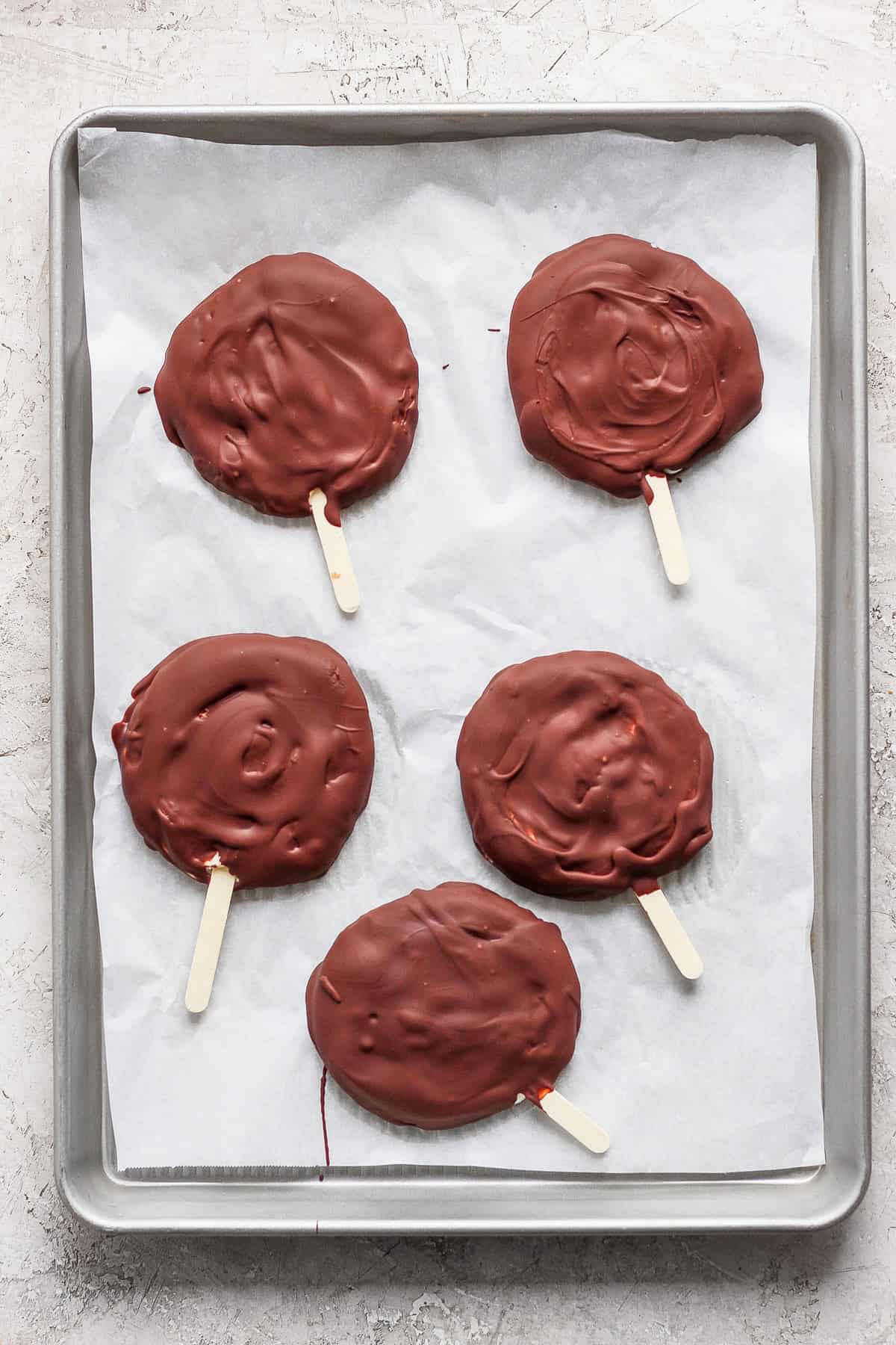 Five chocolate-covered treats on wooden sticks rest on a parchment-lined baking sheet.