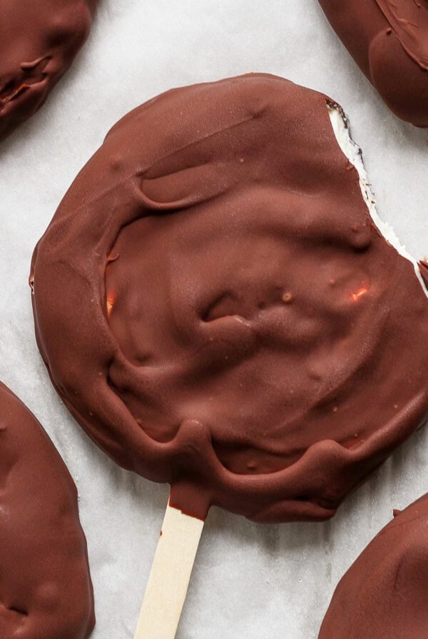 Close-up of a chocolate-covered round ice cream treat on a stick with a bite taken out of the edge, resting on parchment paper along with other similar treats.