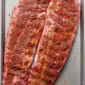 Two slabs of seasoned raw pork ribs are placed on a large rectangular baking sheet, ready for cooking.