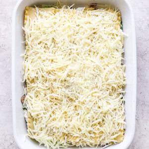 A rectangular white baking dish filled with unbaked lasagna, topped with a layer of shredded cheese, placed on a light-colored surface.