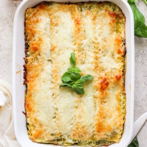 A baked lasagna in a white rectangular dish, topped with melted cheese and garnished with fresh basil leaves. A wooden pepper grinder and fresh basil are visible in the background.