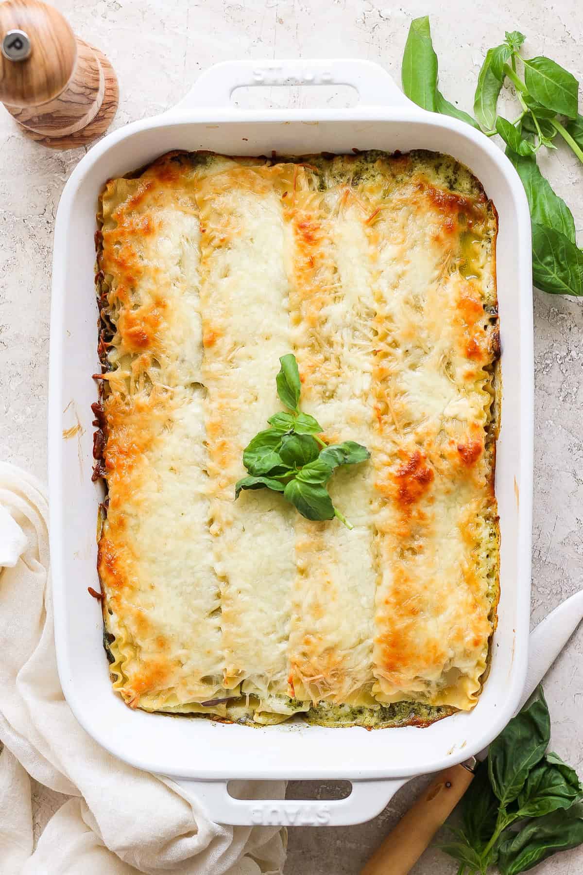 A baked lasagna in a white rectangular dish, topped with melted cheese and garnished with fresh basil leaves. A wooden pepper grinder and fresh basil are visible in the background.