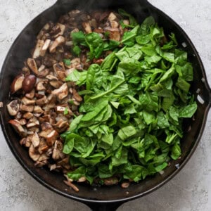 A cast-iron skillet filled with chopped mushrooms on one side and fresh spinach on the other, set on a light-colored surface.