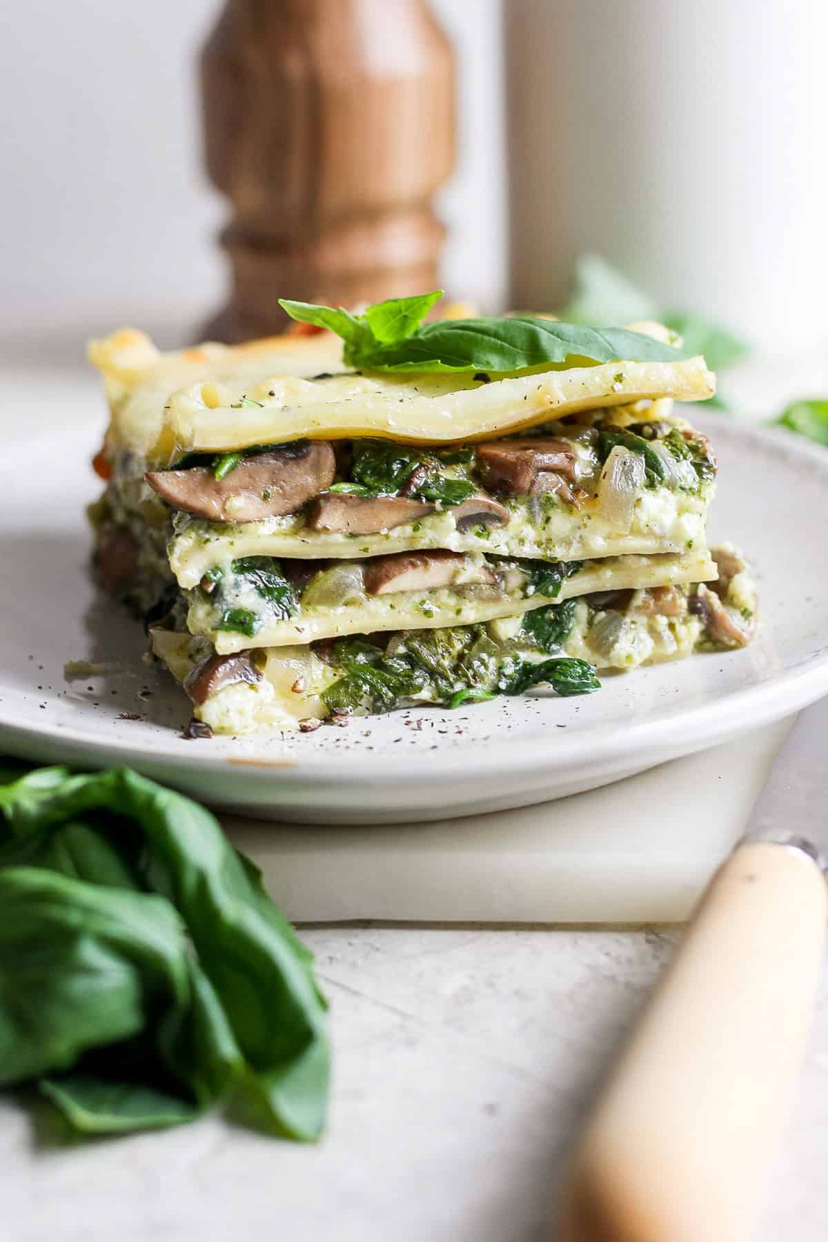 A slice of vegetarian lasagna with visible layers of pasta, spinach, mushrooms, and cheese on a plate, garnished with fresh basil leaves.