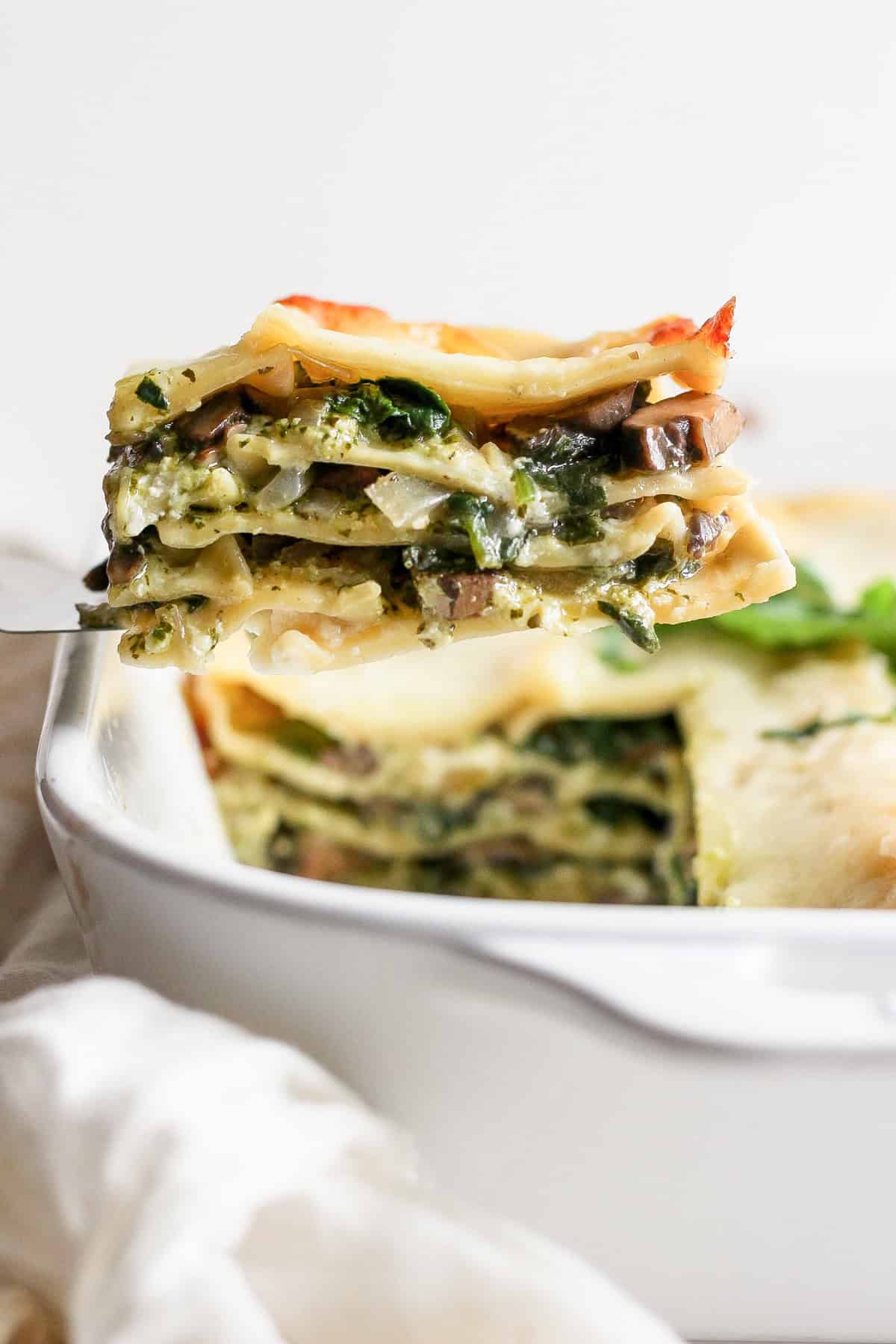 A close-up of a slice of vegetable lasagna being lifted from a white baking dish. The lasagna has layers of pasta, vegetables, and melted cheese.