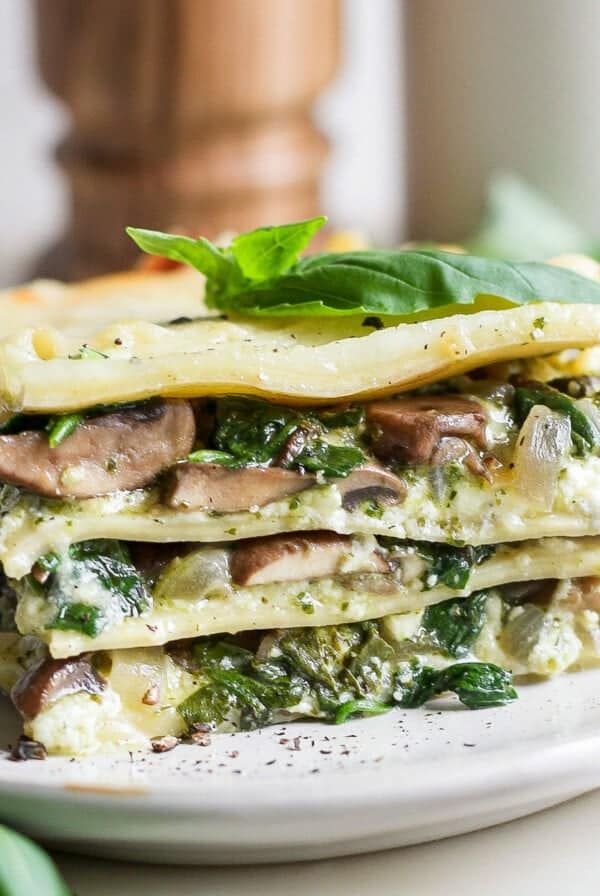 A slice of vegetable lasagna with visible layers of spinach, mushrooms, and cheese on a white plate, garnished with fresh basil leaves.