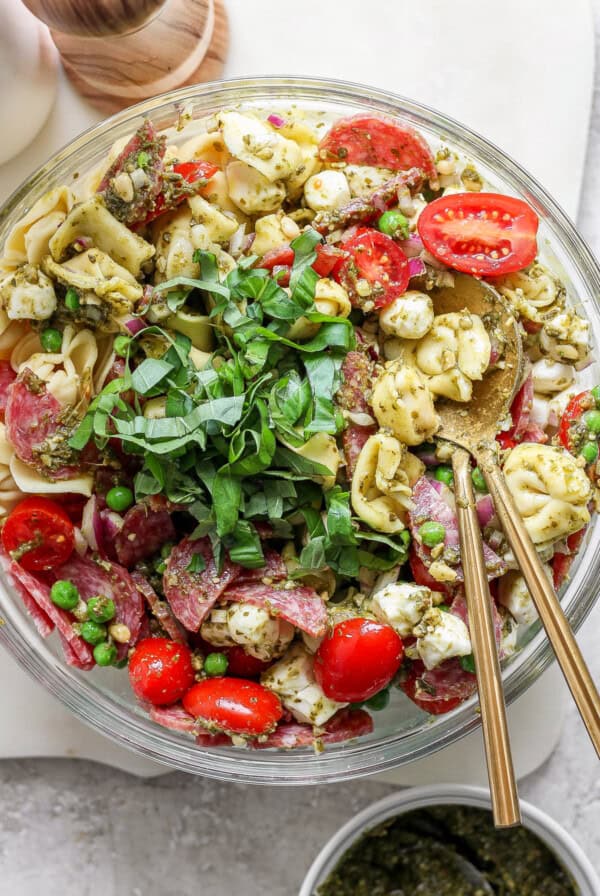 A bowl of tortellini pasta salad with cherry tomatoes, mozzarella, peas, salami, and greens, served with pesto and two spoons.