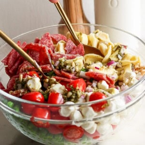 A glass bowl containing ingredients like cherry tomatoes, small mozzarella balls, salami slices, and pasta being mixed with spoons. A bottle and wooden salt grinder are in the background.