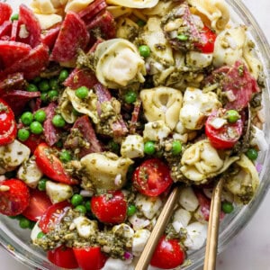 A close-up of a pasta salad with tortellini, cherry tomatoes, peas, mozzarella balls, salami, and green pesto sauce, served in a glass bowl with metal utensils.