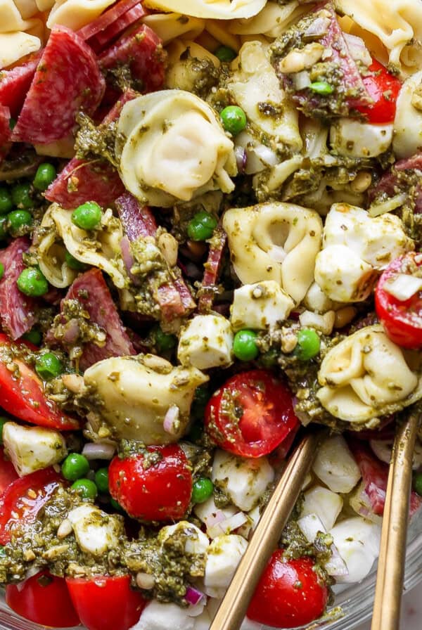 A close-up of a pasta salad with tortellini, cherry tomatoes, peas, mozzarella balls, salami, and green pesto sauce, served in a glass bowl with metal utensils.