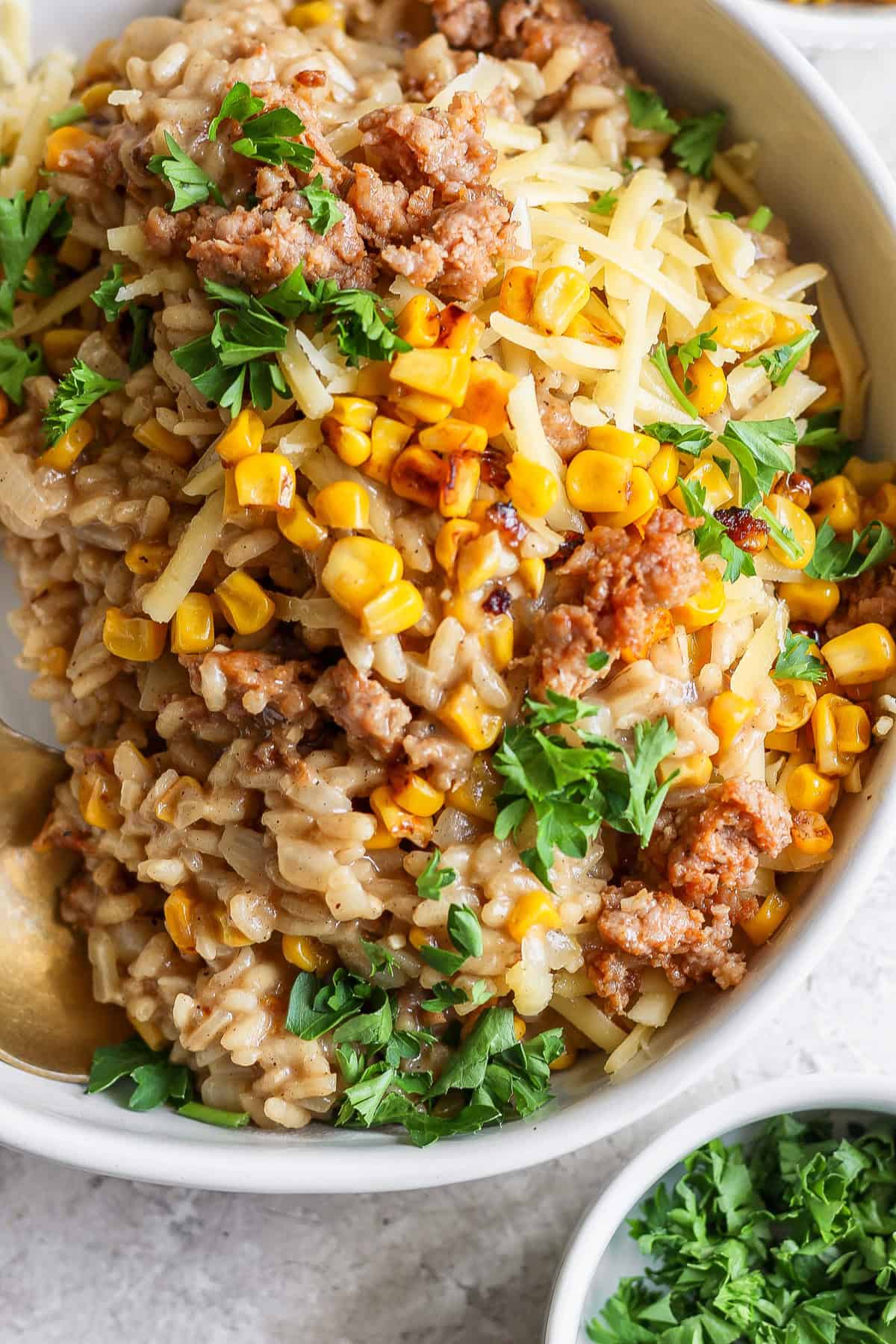 A close-up of a bowl of risotto topped with cooked minced meat, corn kernels, shredded cheese, and fresh parsley, accompanied by a small dish of chopped parsley in the background.