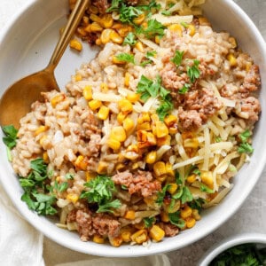 A bowl of barley risotto topped with corn, ground meat, shredded cheese, and garnished with fresh parsley. A gold spoon is placed on the side of the bowl. A small bowl of corn is in the background.