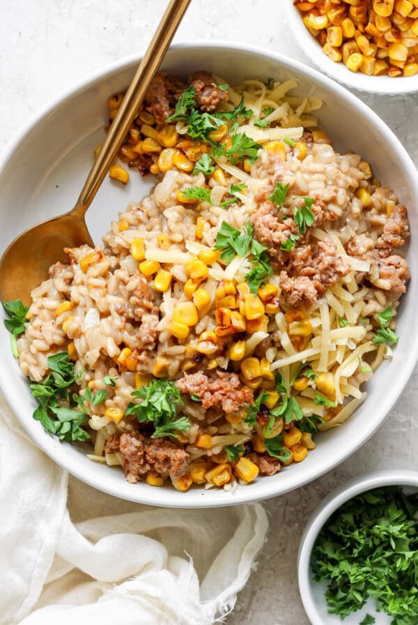 A bowl of barley risotto topped with corn, ground meat, shredded cheese, and garnished with fresh parsley. A gold spoon is placed on the side of the bowl. A small bowl of corn is in the background.