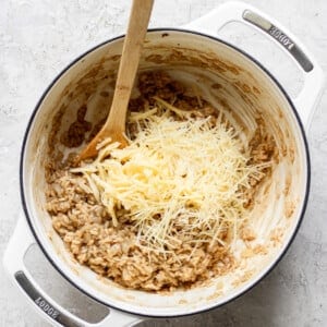 A wooden spoon rests in a white pot containing risotto, with grated cheese on top, ready to be stirred in.