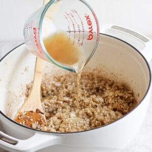 A person pours broth from a measuring cup into a white pot containing sautéed onions, stirring with a wooden spoon.