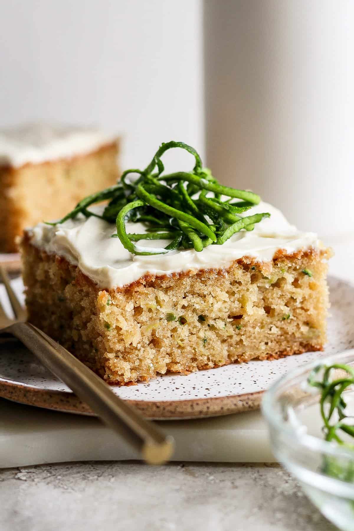 A slice of frosted zucchini cake topped with thin slices of zucchini on a plate with a fork.