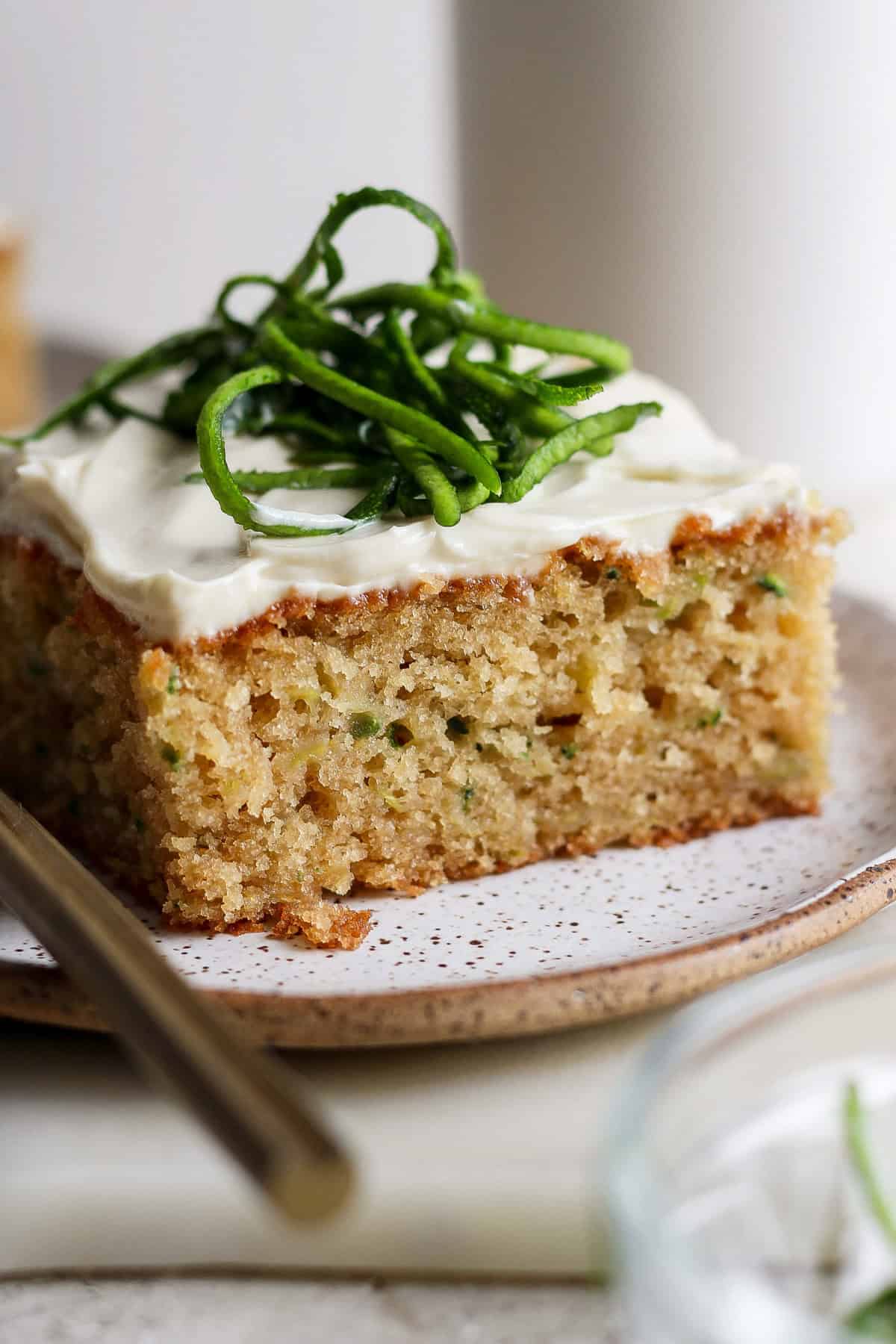 A slice of cake with white frosting and green garnish on a speckled plate, accompanied by a fork.