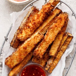 A basket filled with golden brown, crispy breaded vegetable sticks, accompanied by a small bowl of red dipping sauce on the side.