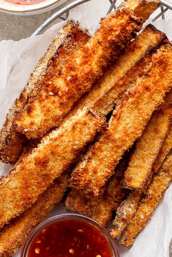 A basket filled with golden brown, crispy breaded vegetable sticks, accompanied by a small bowl of red dipping sauce on the side.