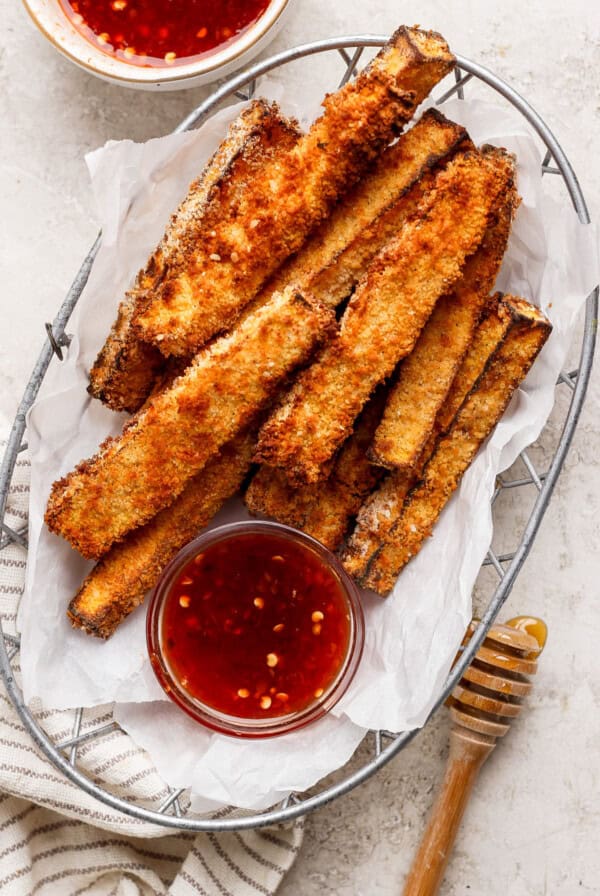 A basket of crispy, breaded zucchini fries served with two small bowls of red dipping sauce and a wooden honey dipper on a striped cloth.