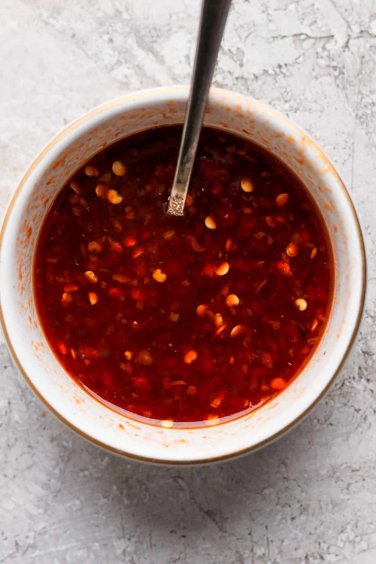 A white bowl filled with a red chili sauce containing visible chili flakes, with a metal spoon resting inside the bowl. The bowl is placed on a textured light gray surface.