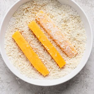 Three breaded cheese sticks are positioned in a bowl filled with breadcrumbs on a textured white surface.