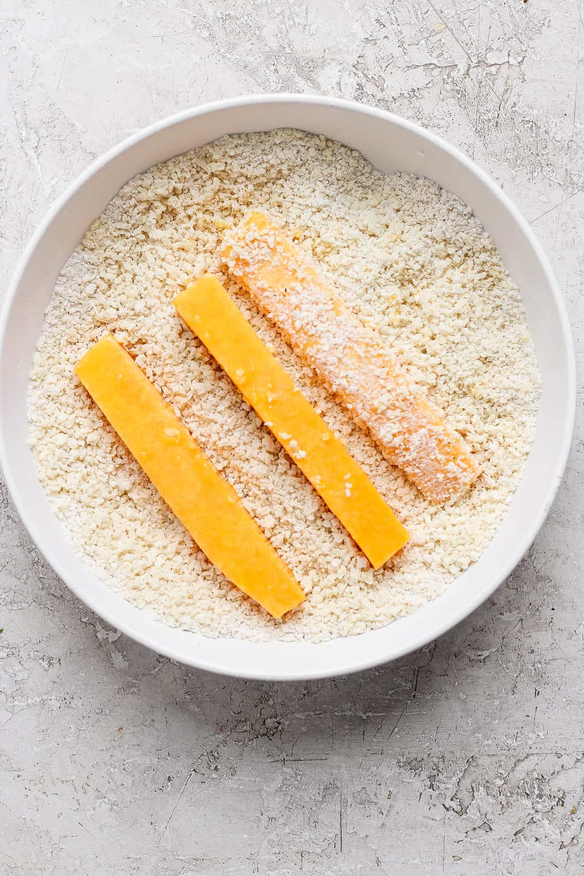 Three breaded cheese sticks are positioned in a bowl filled with breadcrumbs on a textured white surface.