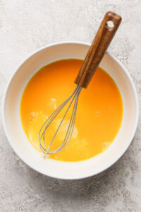 A bowl filled with beaten eggs, with a whisk resting in it on a textured surface.