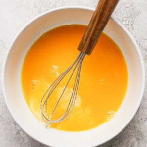 A bowl filled with beaten eggs, with a whisk resting in it on a textured surface.