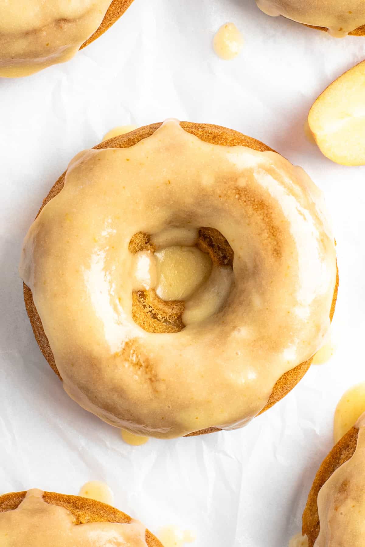 Close-up of a glazed donut with a smooth, shiny coating on a white surface. The donut has a hole in the center with some exposed texture. Another donut is partially visible in the background.