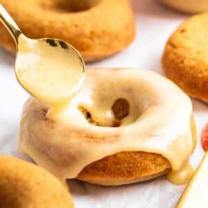 A spoon drizzles glaze over a baked donut with more donuts and a sliced apple in the background.