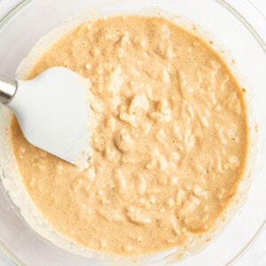 A glass bowl containing a thick, beige batter mixture with a white spatula resting on the side.